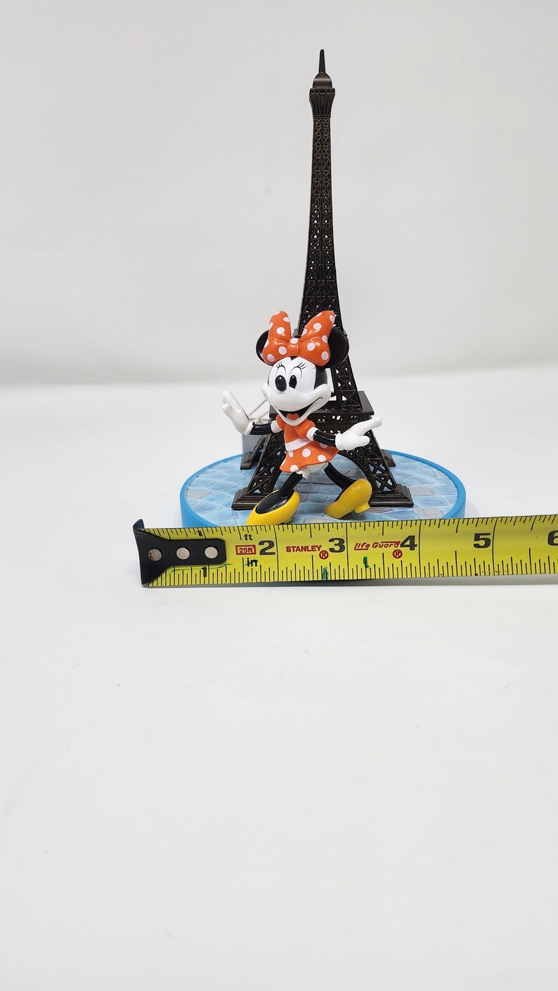 Pu&ograve; includere: Una statuetta in miniatura della Torre Eiffel con il personaggio di Minnie Mouse, che indossa un vestito arancione a pois bianchi e un fiocco abbinato. Un metro a nastro giallo mostra i centimetri.