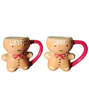 Op de afbeelding: Twee mokken in de vorm van een peperkoeken mannetje. De mokken zijn bruin met witte glazuurdetails, rode strikjes en rode handvatten. De mokken hebben een lachend gezicht en zwarte stippen als knopen.
