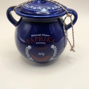 May include: A cobalt blue ceramic paprika jar with a lid and two handles. The jar has text that reads "Kalocsai Fűszer Paprika őrlemény Csípős 50g" in white lettering. A decorative cord secures the lid.