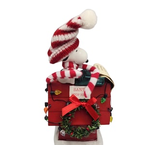 May include: A Snoopy Christmas decoration featuring Snoopy wearing a red and white striped hat and scarf. Snoopy is perched atop a red mailbox with the words "SANTA STOP HERE" and a small wreath. The mailbox is decorated with string lights.