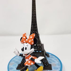 Pu&ograve; includere: Una statuina di Minnie Mouse con un vestito e fiocco a pois arancioni, con una Torre Eiffel in miniatura sullo sfondo. La base blu riporta la scritta "Disneyland Paris". La Torre Eiffel &egrave; alta circa 13 cm.