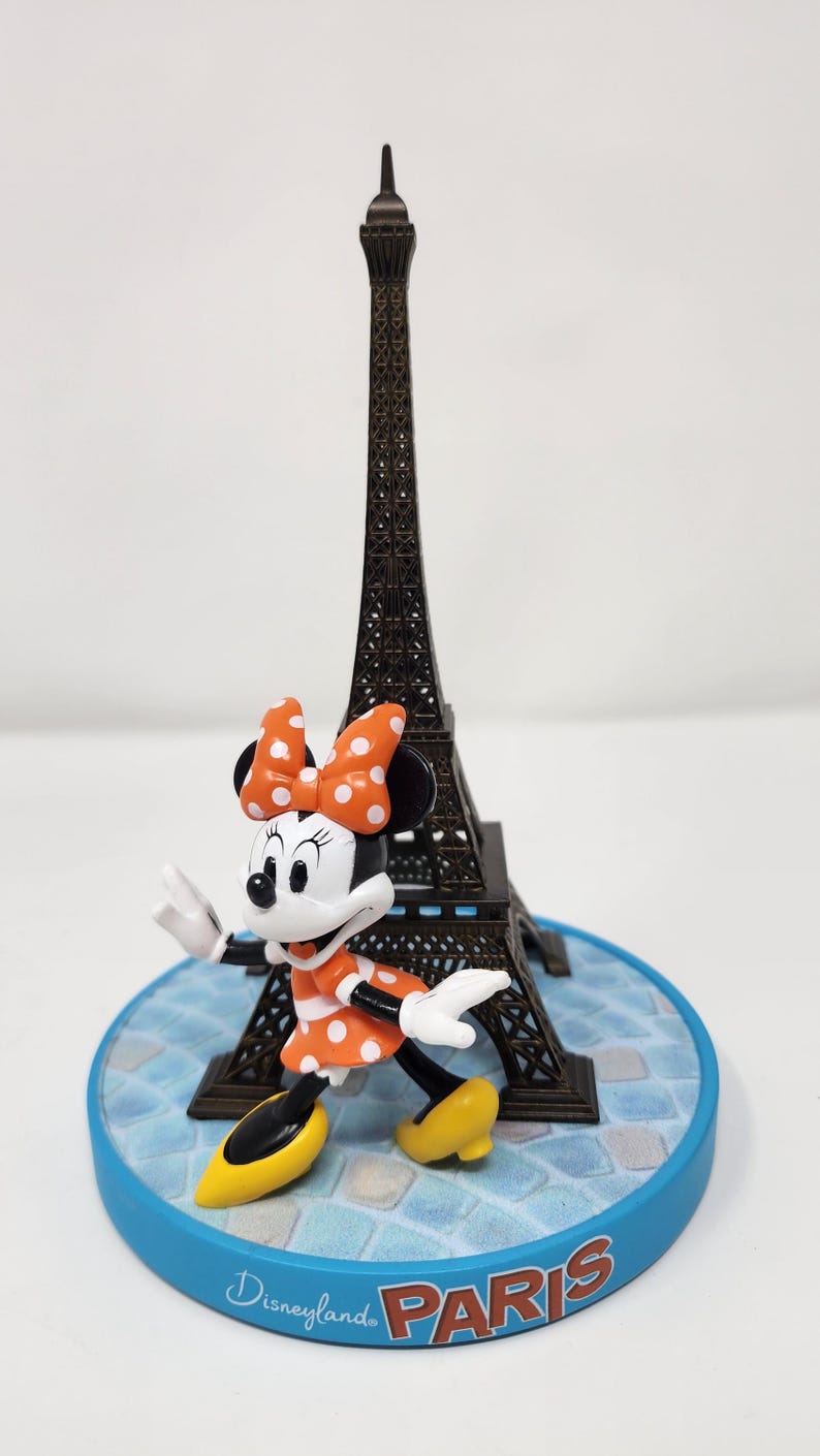 Pu&ograve; includere: Una statuina di Minnie Mouse con un vestito e fiocco a pois arancioni, con una Torre Eiffel in miniatura sullo sfondo. La base blu riporta la scritta "Disneyland Paris". La Torre Eiffel &egrave; alta circa 13 cm.