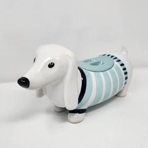 Op de afbeelding: Een witte keramische teckelvormige container met een lichtblauw en wit gestreept ontwerp. De hond heeft zwarte ogen en een zwarte neus. De container heeft een deksel met een handvat. De hond staat op vier poten.