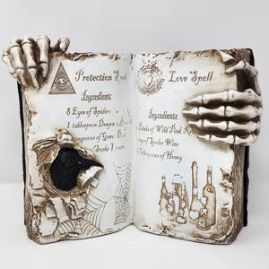 Può includere: Un libro aperto decorativo dall'aspetto invecchiato. Le pagine presentano istruzioni di incantesimi scritte a mano, tenute aperte da mani scheletriche. La pagina sinistra ha un buco e un disegno a ragnatela. La pagina destra ha illustrazioni di bottiglie.