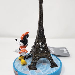 Pu&ograve; includere: Una statuetta in miniatura della Torre Eiffel in bronzo accanto a una statuetta di Minnie Mouse. Minnie indossa un vestito arancione a pois bianchi e un fiocco abbinato. Le statuette sono su una base blu con la scritta "Paris".