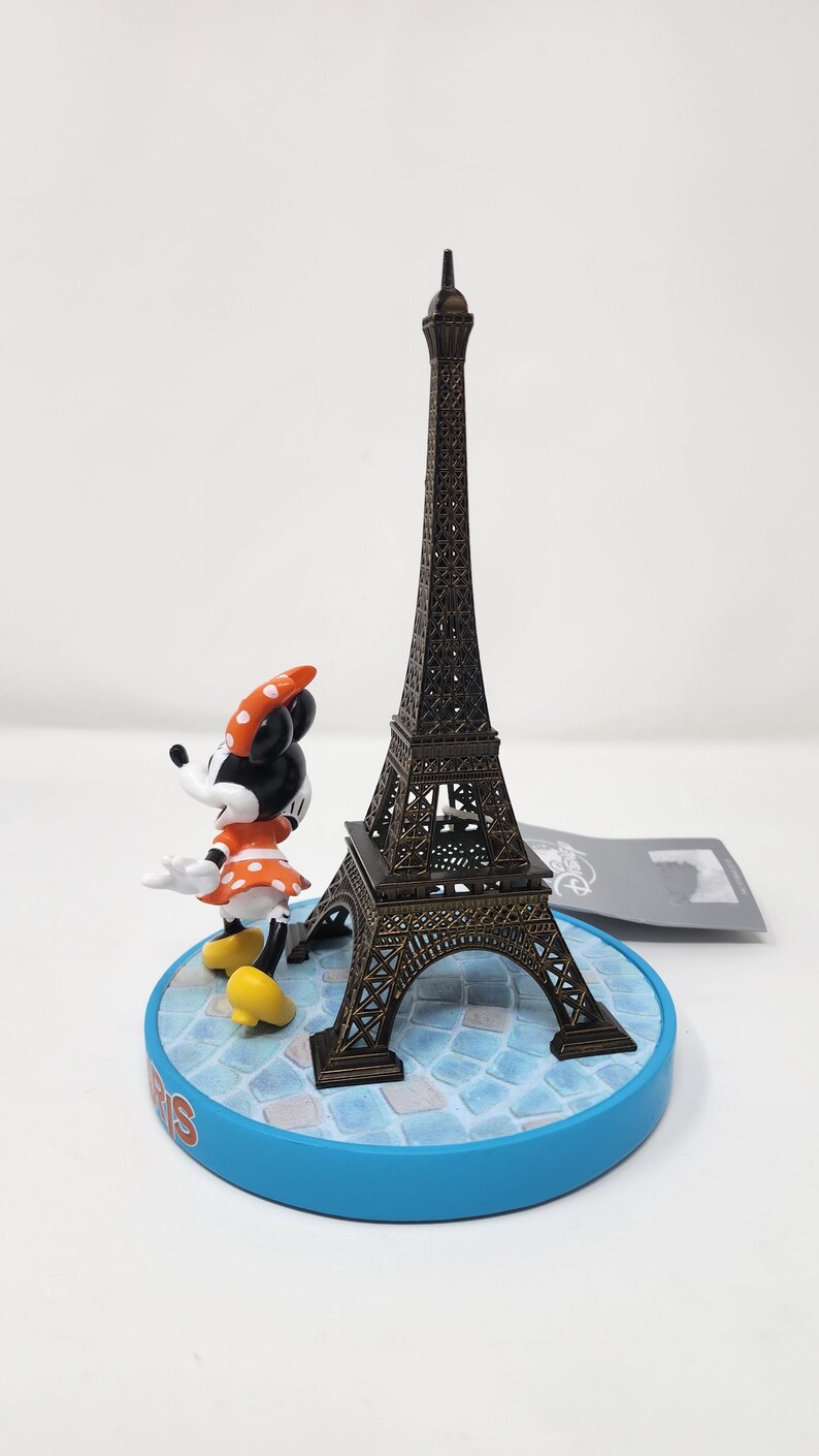 Pu&ograve; includere: Una statuetta in miniatura della Torre Eiffel in bronzo accanto a una statuetta di Minnie Mouse. Minnie indossa un vestito arancione a pois bianchi e un fiocco abbinato. Le statuette sono su una base blu con la scritta "Paris".