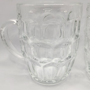 Op de afbeelding: Twee heldere bierpullen van glas met een gestructureerd, paneelontwerp. Elke pul heeft een stevige handgreep en een brede, afgeronde basis. De pullen zijn leeg en lijken schoon te zijn.