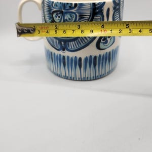 May include: A white ceramic mug with a blue floral design and a handle. The mug is being measured with a tape measure, showing a width of approximately 4 inches.