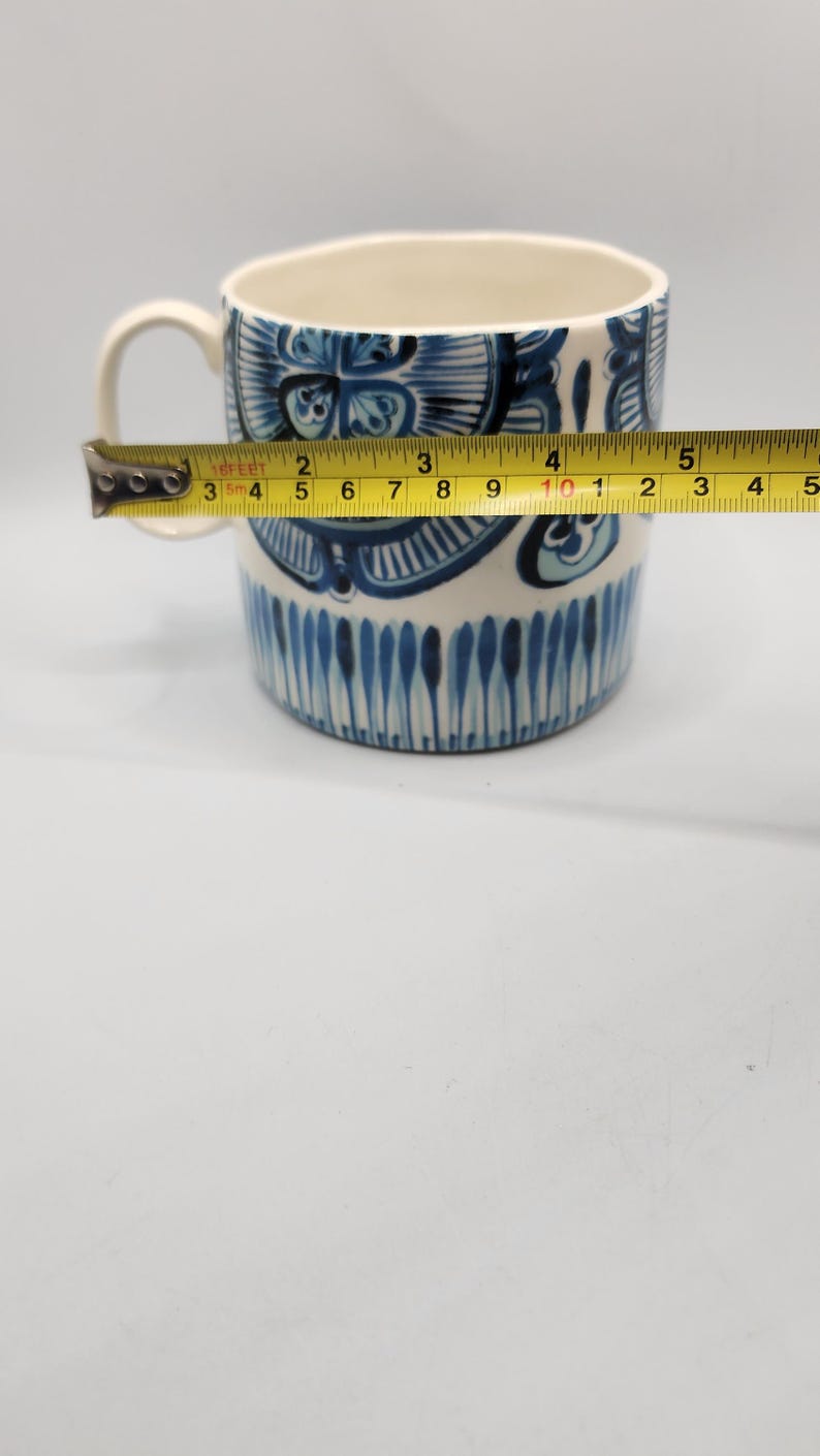 May include: A white ceramic mug with a blue floral design and a handle. The mug is being measured with a tape measure, showing a width of approximately 4 inches.