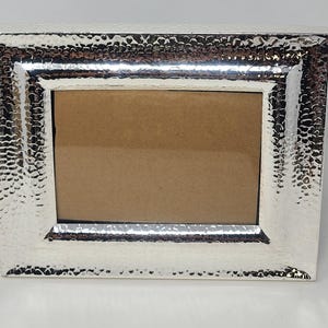 May include: A rectangular picture frame with a metallic silver finish and a distinct hammered texture across the entire surface. The frame features a wide border surrounding a central rectangular opening that contains a plain brown insert.