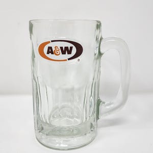 May include: Clear glass A&W root beer mug with a handle. The mug has a rectangular shape with vertical ridges and the A&W logo in brown and orange. The mug is empty and sits on a white surface.