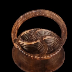 May include: A handcrafted wooden basket with a handle. The basket is made of multiple layers of wood, creating a unique, layered design. The handle and rim feature decorative carvings. The basket is brown and sits on a round base.