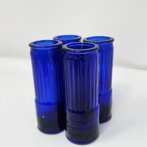 May include: Four cobalt blue glass shot glasses. Each glass has a cylindrical shape with vertical ridges and a slightly flared rim. The base of each glass has embossed text. The glasses are arranged on a white surface.