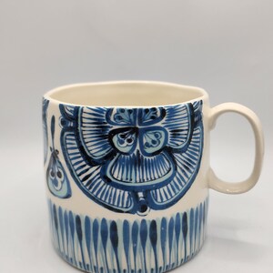 May include: A ceramic mug with a cream-colored interior and handle. The exterior features a hand-painted design in shades of blue, including floral and vertical stripe patterns. The mug is suitable for hot or cold beverages.