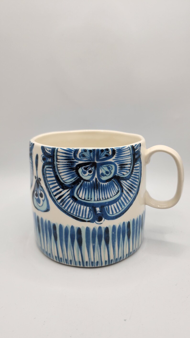 May include: A ceramic mug with a cream-colored interior and handle. The exterior features a hand-painted design in shades of blue, including floral and vertical stripe patterns. The mug is suitable for hot or cold beverages.