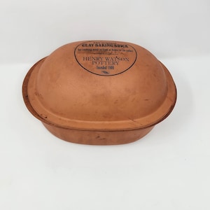 Puede incluir: Un ladrillo de hornear de arcilla terracota ovalado con tapa abovedada. La parte superior presenta texto negro que dice: The original CLAY BAKING BRICK for cooking meat or fowl at home or on safari HENRY WATSON POTTERY founded 1800. Superficie marrón mate.