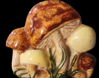May include: A ceramic mushroom sculpture with a cluster of mushrooms in shades of brown, yellow, and beige. The mushroom caps have a textured appearance, and green blades of grass are visible. The sculpture is set against a black background.
