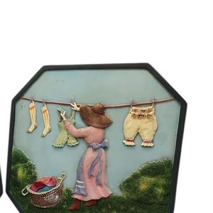 Può includere: Scultura a rilievo incorniciata raffigurante una donna che stende il bucato. La donna indossa un vestito rosa e un cappello marrone. La scena comprende calzini, un vestito e pantaloni ad asciugare, con un cesto di vestiti in primo piano.