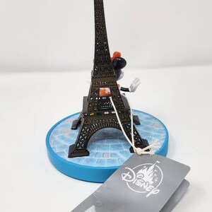 Pu&ograve; includere: Una statuetta in miniatura della Torre Eiffel con un personaggio Disney. La torre in metallo marrone poggia su una base blu con un motivo a mosaico. Un'etichetta Disney &egrave; appesa alla statuetta. La statuetta &egrave; alta circa 15 cm.
