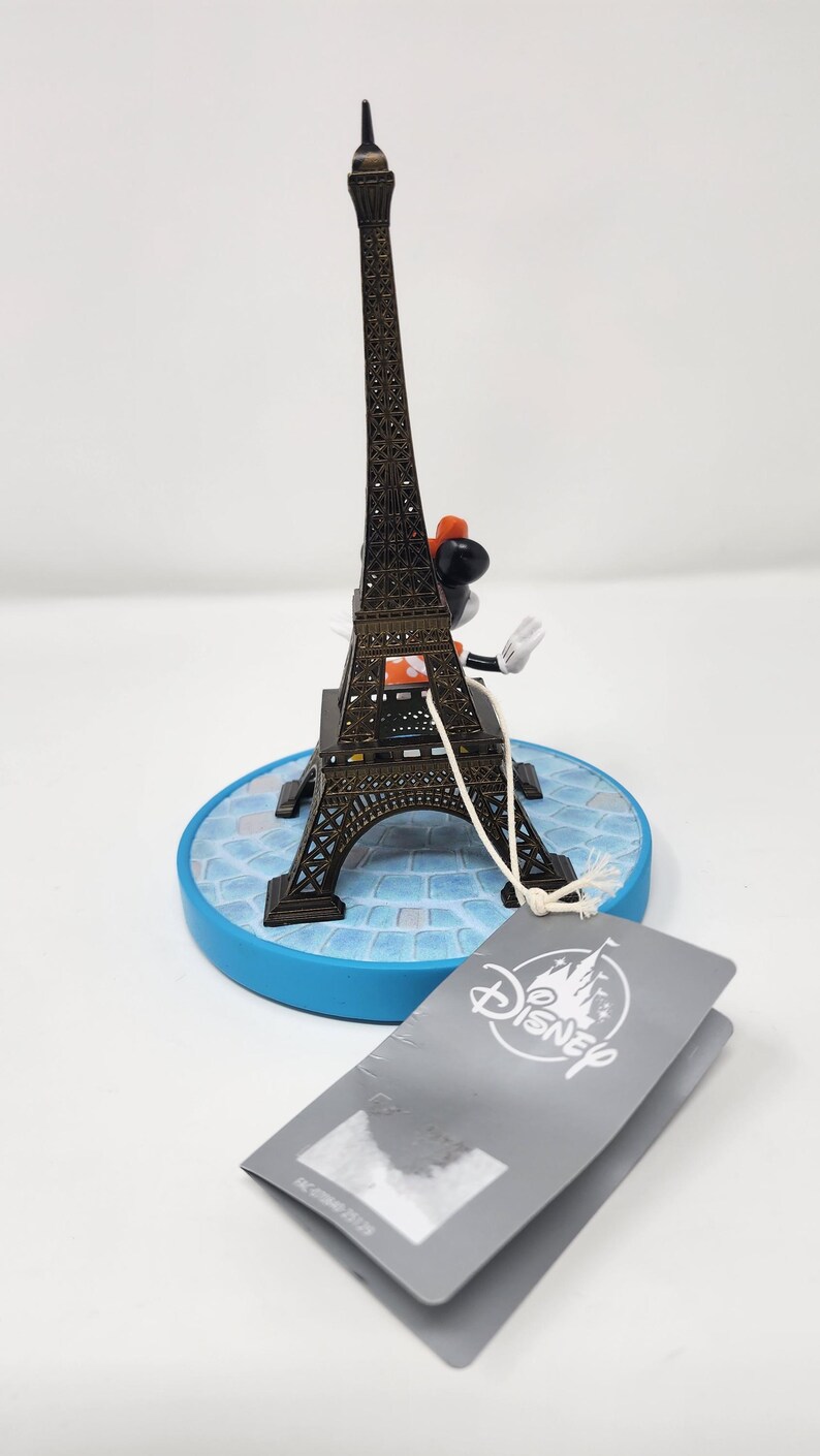 Pu&ograve; includere: Una statuetta in miniatura della Torre Eiffel con un personaggio Disney. La torre in metallo marrone poggia su una base blu con un motivo a mosaico. Un'etichetta Disney &egrave; appesa alla statuetta. La statuetta &egrave; alta circa 15 cm.