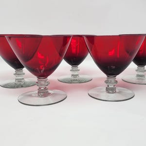 Könnte beinhalten: Ein Set aus fünf rubinroten Cocktailgläsern mit breiten, V-förmigen Kelchen und klaren, mehrstufigen Stielen. Jedes Glas hat eine glatte, durchscheinende rote Oberfläche, die in einen klaren, kreisförmigen Fuß übergeht und einheitlich wirkt.