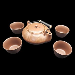 May include: A brown ceramic tea set with a teapot and four matching teacups. The teapot has a black handle and a textured surface. The teacups are small and round, with a simple design. The set is displayed against a black background.