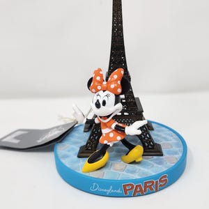 Pu&ograve; includere: Una statuina di Minnie Mouse con un vestito arancione a pois, davanti a una Torre Eiffel in miniatura. La base blu reca la scritta "Disneyland Paris". Minnie indossa scarpe gialle e un fiocco abbinato.