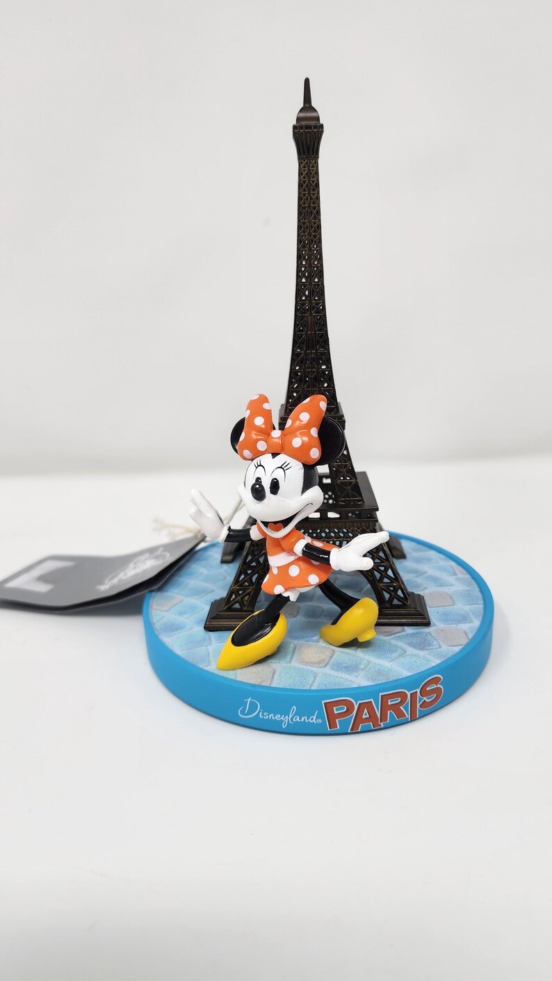 Pu&ograve; includere: Una statuina di Minnie Mouse con un vestito arancione a pois, davanti a una Torre Eiffel in miniatura. La base blu reca la scritta "Disneyland Paris". Minnie indossa scarpe gialle e un fiocco abbinato.