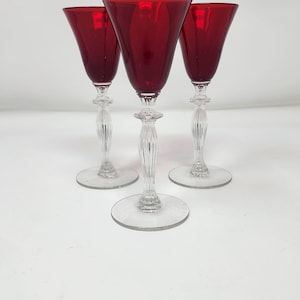 May include: Three elegant red wine glasses with clear, ornate stems and round bases. The ruby-colored bowls contrast with the clear stems, creating a sophisticated look. The glasses are arranged on a white surface.