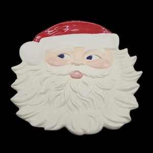 May include: A ceramic Santa Claus face with a red hat and white beard. The face has rosy cheeks, blue eyes, and a pink mouth. The beard is full and textured, and the hat has a white trim.