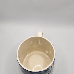 May include: Cream-colored ceramic mug with a blue floral pattern around the exterior. The mug has a small, rectangular handle and a smooth, glossy finish. The interior is a lighter cream color.