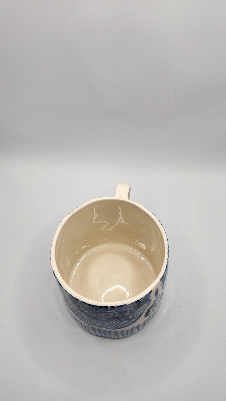 May include: Cream-colored ceramic mug with a blue floral pattern around the exterior. The mug has a small, rectangular handle and a smooth, glossy finish. The interior is a lighter cream color.