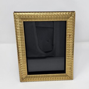 May include: A rectangular picture frame with a metallic gold finish. The frame features a repeating embossed pattern along the border, consisting of vertical lines and small dots. The centre of the frame contains a solid black rectangular insert.