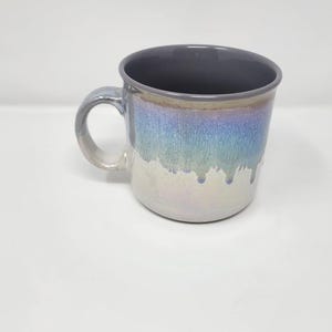 May include: A ceramic mug featuring a dark gray interior and a handle. The exterior displays a reactive glaze with a gradient transition from a metallic blue and purple iridescent top section to a glossy, light cream-colored base with drips.