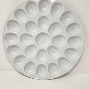 Puede incluir: Una fuente redonda blanca con 28 huecos en forma de huevo. La fuente es de cerámica y tiene un acabado brillante. El diseño es simétrico, con una hendidura central rodeada de anillos concéntricos.