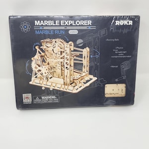 May include: A box for a wooden marble run kit, labeled "Marble Explorer" and "Marble Run LG503." The box features a diagram of the assembled model, with text indicating "Running Balls" and "Manual." A warning about choking hazards is also present.