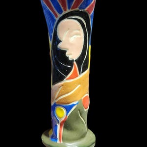 May include: A tall, ceramic vase with a stylized portrait of a woman. The vase features a color palette of blue, red, yellow, orange, and black. The base is green, and the background is black. The vase is approximately 8 inches tall.