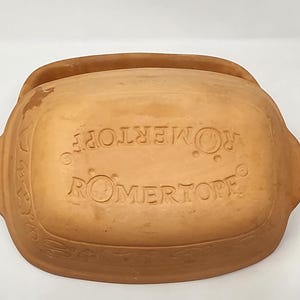 May include: A rectangular terracotta Romertopf clay baker with a lid. The top of the lid features the embossed text ROMERTOPF and decorative relief carvings of various vegetables and leaves along the side edges, with integrated side handles.