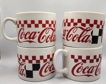 Op de afbeelding: Vier witte keramische Coca-Cola mokken. Elke mok heeft het Coca-Cola logo in rood, met een rood en zwart geblokt patroon. De mokken hebben een gebogen handvat en zijn per paar gestapeld.