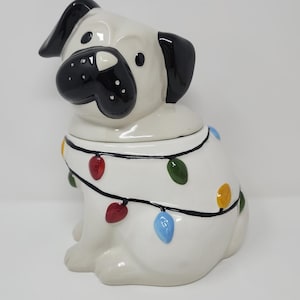 May include: A ceramic pug-shaped cookie jar. The white pug has black ears and a black nose. It is decorated with a string of colourful Christmas lights. The jar is designed to look like the pug is wearing the lights.