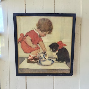 May include: A square wall art print featuring a child with curly brown hair wearing a red dress and striped socks, pouring milk from a blue and white pitcher into a bowl for a black and white kitten with a red bow. The artist name Jessie Willcox Smith is visible.