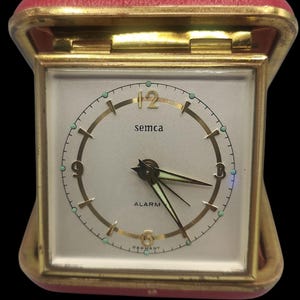 May include: A vintage Semca alarm clock in a red and gold case. The clock face is white with gold hands and numerals. The word "semca" is printed on the face, and the word "alarm" is printed below the hands. The clock is marked "Germany".