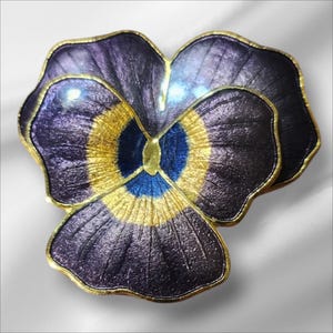 May include: A detailed, close-up view of a purple, gold, and blue pansy flower brooch. The brooch has a gold-colored trim and a textured surface. The flower's petals are a deep purple, with a gold and blue center.