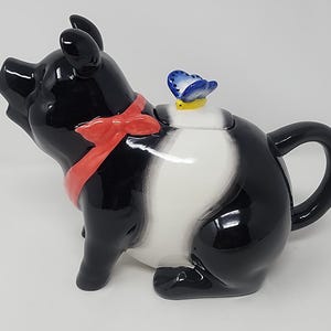 Peut inclure: Une théière en céramique en forme de cochon avec une finition noire et blanche brillante. Le cochon porte un ruban rouge vif autour du cou et un petit papillon bleu et jaune est posé sur le couvercle. Elle possède une anse courbée.