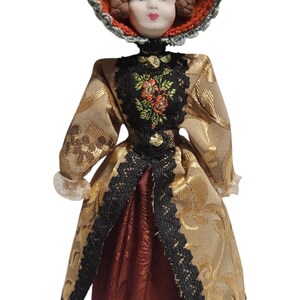 May include: A vintage porcelain doll with a detailed face, wearing a gold brocade dress with black lace trim and a matching hat. The doll's dress features a floral embroidered bodice and a burgundy skirt.