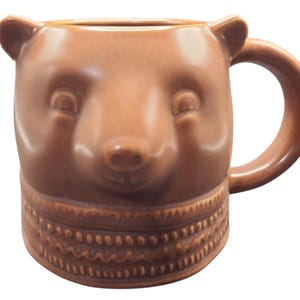 Op de afbeelding: Bruine keramische mok in de vorm van een berenkop. De mok heeft een gedetailleerd berengezicht met kleine oren en een gestructureerde band rond de basis. De handgreep is een eenvoudige lus.