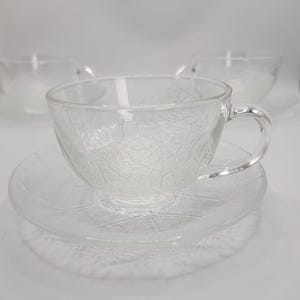 May include: A clear glass teacup and saucer set. The teacup has a rounded shape with a handle and sits on a matching saucer. Both pieces feature a leaf pattern. Suitable for serving tea or coffee.