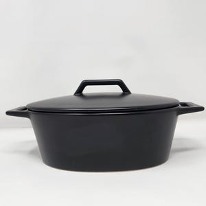 May include: A matte black oval Dutch oven with a matching lid. The pot features two integrated side handles and a central handle on the lid. The exterior surface is smooth and uniform in color, showcasing a wide, shallow basin design.