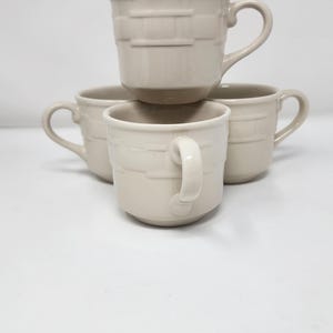 Puede incluir: Cuatro tazas de café de cerámica beige apiladas sobre una superficie blanca. Las tazas tienen un diseño sencillo con un asa curvada y un patrón de bandas horizontales texturizadas.