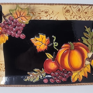 May include: A rectangular ceramic serving tray with rounded corners and two side handles. The tray features a glossy black centre decorated with a raised, three-dimensional painted design of a pumpkin, red apple, grapes, and autumn leaves.